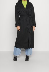 Trench-coat noir avec boutons croisés et taille ceinturée. Associé à un haut vert et un jean à jambes larges avec ourlet effiloché. Bottines noires.