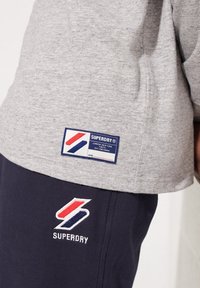 Graues kurzärmeliges Hemd mit einem blauen und roten Superdry-Logo-Tag am Saum, kombiniert mit marineblauen Hosen mit einem gestickten Superdry-Logo.