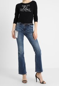 Top nero a maniche lunghe con scritte metalliche, abbinato a jeans svasati blu. Tacchi neri aperti e una pochette argentata completano il look.