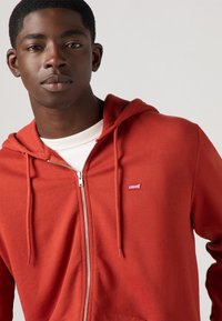Felpa rossa con zip e cappuccio, maglietta bianca sotto e piccolo logo Levi's sul petto. Morbido al tatto e design casual.