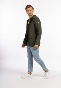 Jachetă impermeabilă de culoare verde măslin cu glugă, închidere cu fermoar și două buzunare laterale, combinată cu blugi albaștri deschisi și adidași albi.