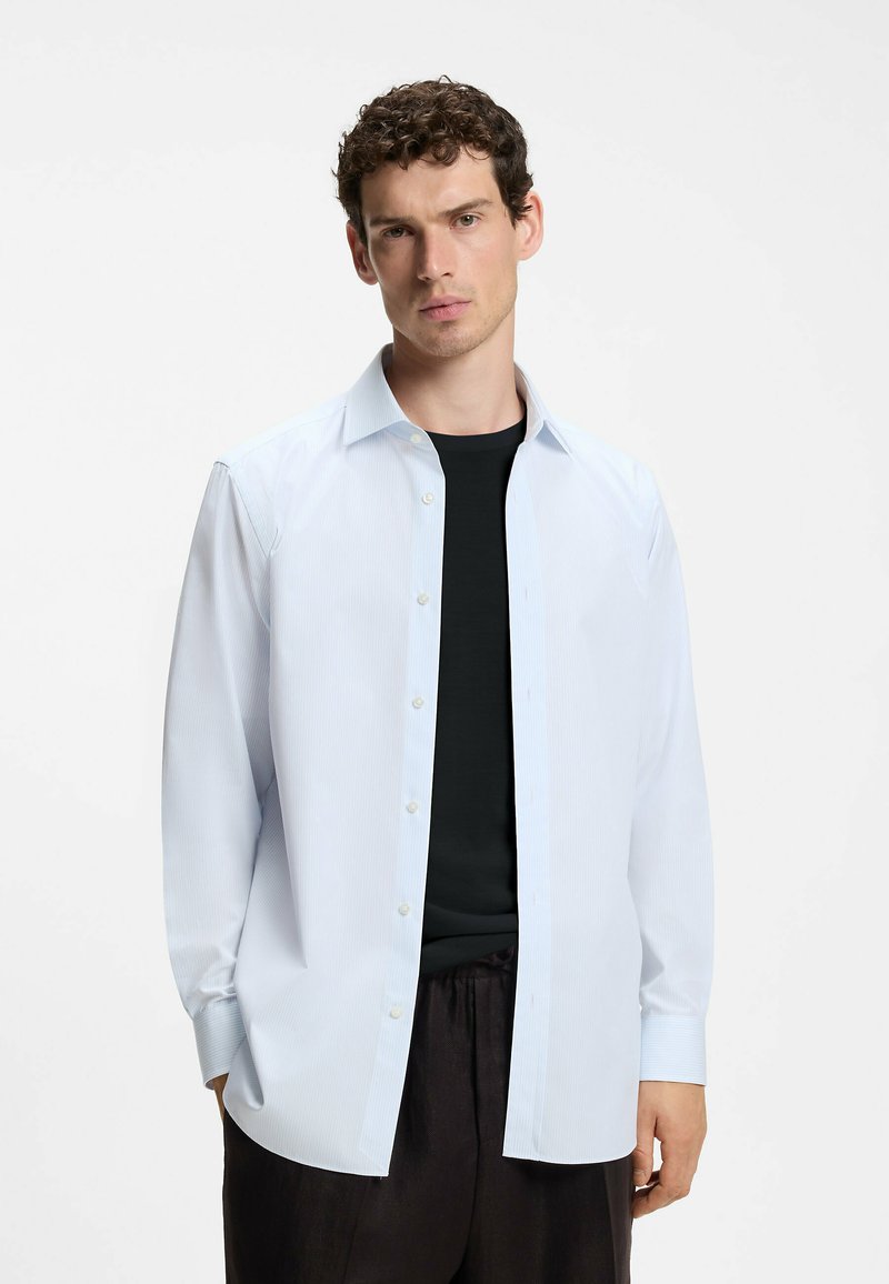 Homme aux cheveux courts et bouclés portant une chemise bleu clair ouverte à boutons sur un t-shirt noir et un pantalon noir, sur un fond uni.