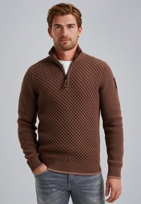 Bruine gebreide pullovertrui met een gestructureerd ruitpatroon, kraag met rits tot een kwart, geribbelde manchetten en zoom, gecombineerd met een wit shirt daaronder.
