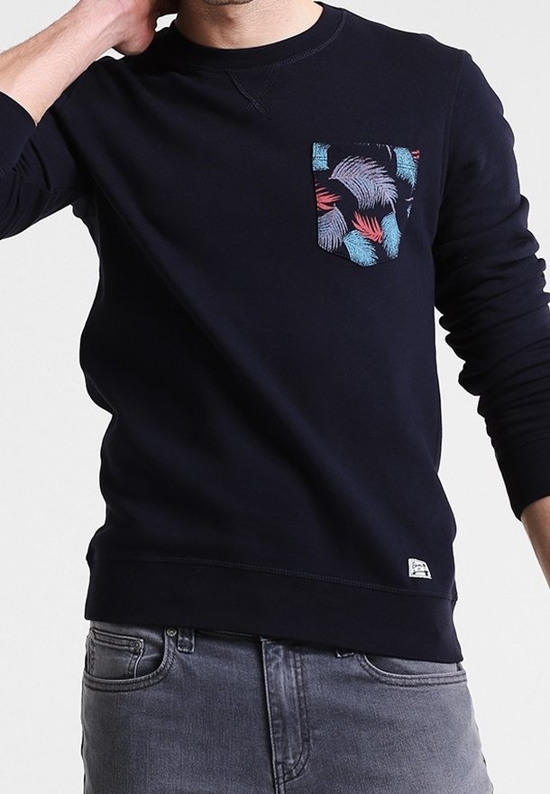 Mann, der ein schwarzes langärmliges Hemd mit einer bunten Feder-Muster Brusttasche und heller grauer Jeans trägt.