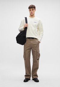 Langärmliges cremefarbenes Hemd mit blauer Stickerei, braune Cargo-Hosen mit Seitentaschen und schwarze Schuhe; das Model trägt eine schwarze Umhängetasche.