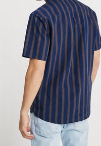 Camisa de manga corta en azul marino con rayas verticales en marrón, con cuello clásico y un plisado simple en la parte trasera, confeccionada en tela ligera.