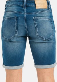 Pantalones cortos de mezclilla en azul medio, con dobladillo enrollado, diseño de cinco bolsillos y una etiqueta de cuero marrón en la cintura para la marca.