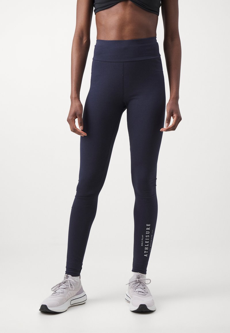 Navy high-waisted leggings made of stretchy fabric, featuring a smooth texture and printed text along the lower leg. Paired with gray sneakers.