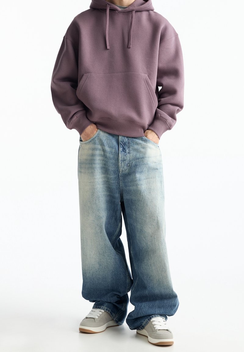 Personne portant un sweat à capuche mauve, un jean bleu délavé ample et des baskets blanches, debout les mains dans les poches devant un fond blanc.