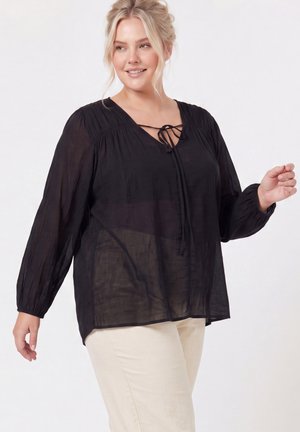Femme souriante aux cheveux blonds portant un chemisier noir à manches longues avec un détail de lien et un pantalon beige sur un fond uni.