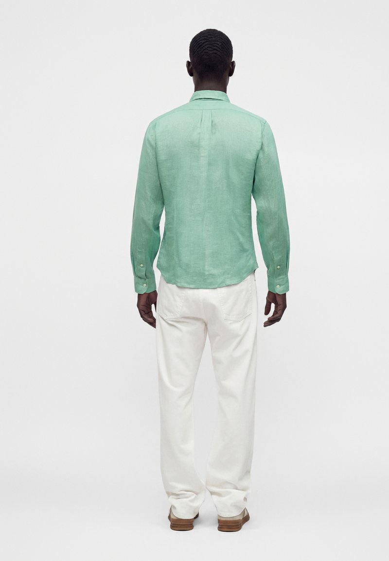 Homme vu de dos, portant une chemise à manches longues vert clair, un pantalon blanc et des chaussures marron sur un fond blanc uni.