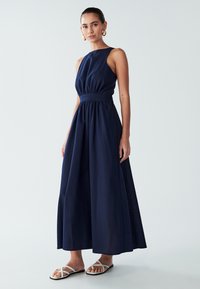 Kvinna klädd i en ärmlös långklänning i marinblått med åtsittande midja och vita sandaletter med remmar, stående mot en ljus, enfärgad bakgrund.