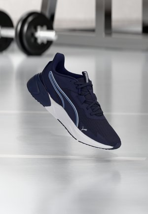 Zapatilla Puma azul marino con suela, cordones y logo blancos, flotando sobre un suelo de gimnasio con pesas al fondo.