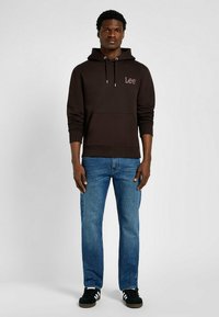 Brązowy hoodie z regulowanym sznurkiem, przednią kieszenią kangurką i subtelnym logo Lee; w komplecie z niebieskimi dżinsami i czarnymi trampkami.
