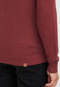 Maglione rosso lavorato a maglia con polsini e orlo a coste, con toppe sui gomiti e un piccolo etichetta logo beige vicino al fondo.