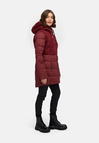 Burgunderfarbene Pufferjacke mit Kapuze, featuring einem kontrastierenden oberen Teil, seitlichen Taschen und quilted Design. Kombiniert mit schwarzen Slim-Fit-Hosen und klobigen Stiefeln.