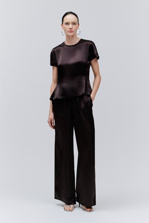 Mujer de pie de frente con una camiseta corta negra brillante, pantalones negros de pierna ancha, pendientes de aro plateados y tacones abiertos plateados.