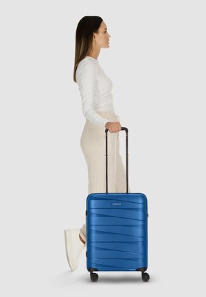 Donna in top bianco e pantaloni beige che cammina con una valigia blu con ruote e manico esteso su uno sfondo neutro.