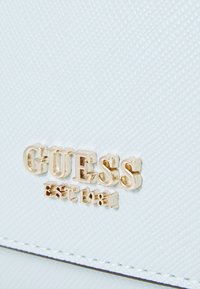 Baltas tekstūruotas rankinukas su aukso "GUESS" logotipu ir „EST 1981“ detale. Lygi apdaila su išryškinta raštu ir kontrastinga siuvinėta.