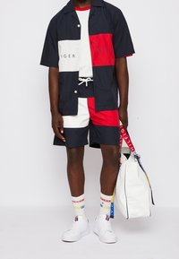 Camicia a maniche corte a blocchi di colore in blu navy, rosso e bianco con testo logo; pantaloncini abbinati; sneakers bianche; borsa bianca di medie dimensioni con tracolle colorate.