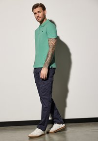 Zelené polo tričko s límečkem, ve spojením s námořnickými cargo kalhotami. Model má na sobě bílé tenisky. Tetování viditelné na pažích. Jednoduchý design a střih.