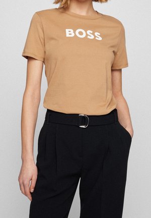 Camiseta de algodón marrón con el texto "BOSS" en blanco, combinada con pantalones negros de cintura alta con una hebilla de cinturón plateada y frente plisado.