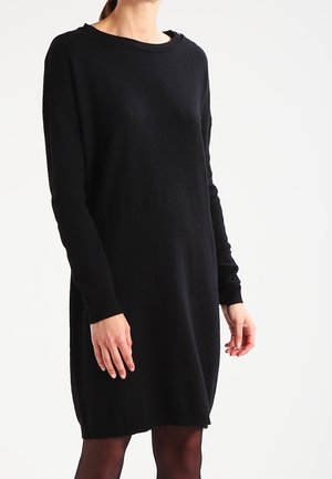 Vrouw draagt een zwart, knielang trui-jurk met lange mouwen en een ronde hals, tegen een effen witte achtergrond.