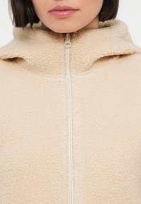 Μπεζ φλις μπουφάν με ψηλό γιακά, που διαθέτει εμπρόσθιο φερμουάρ και απαλό, υφασμάτινο υλικό.