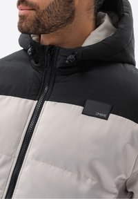 Puffer jacket featuring a color-block design with a black upper section and light gray lower section. Includes a zip closure and hood.