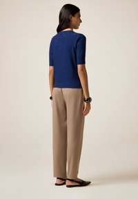 Donna in piedi di lato che indossa un maglione blu a maniche corte con macchie, pantaloni beige, bracciali neri e décolleté neri.