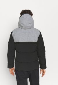 Chaqueta acolchada con un cuerpo negro y una sección superior gris, con capucha y tejido texturizado. El diseño incluye costuras horizontales y una banda con logo.