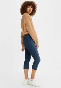 Blauwe denim capri's met een taps toelopende pasvorm en een rafelrand, gecombineerd met een beige oversized sweatshirt en witte sneakers. Zachte textuur overal.