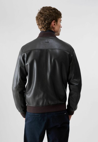 Homme vu de dos portant une veste en cuir noire avec un col côtelé marron, des poignets et une ceinture, sur un fond gris uni.