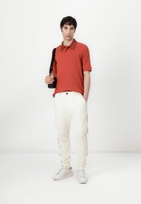 Rød polo skjorte med kontrasterende krage, kombinert med hvite kordfløyelsbukser med svart sidestripe, og hvite sneakers.