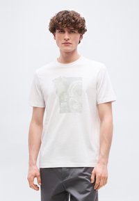 Νέος άνδρας με σγουρά μαλλιά που φοράει ένα λευκό γραφικό t-shirt BOSS και γκρι παντελόνια, στέκεται μπροστά σε ένα απλό φωτεινό φόντο.