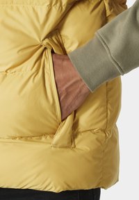 Gelb gefütterte Weste mit strukturiertem Äußeren und Seitentasche, im Vordergrund eine Nahaufnahme einer Hand, die in die Tasche greift, vor einem neutralen Hintergrund.