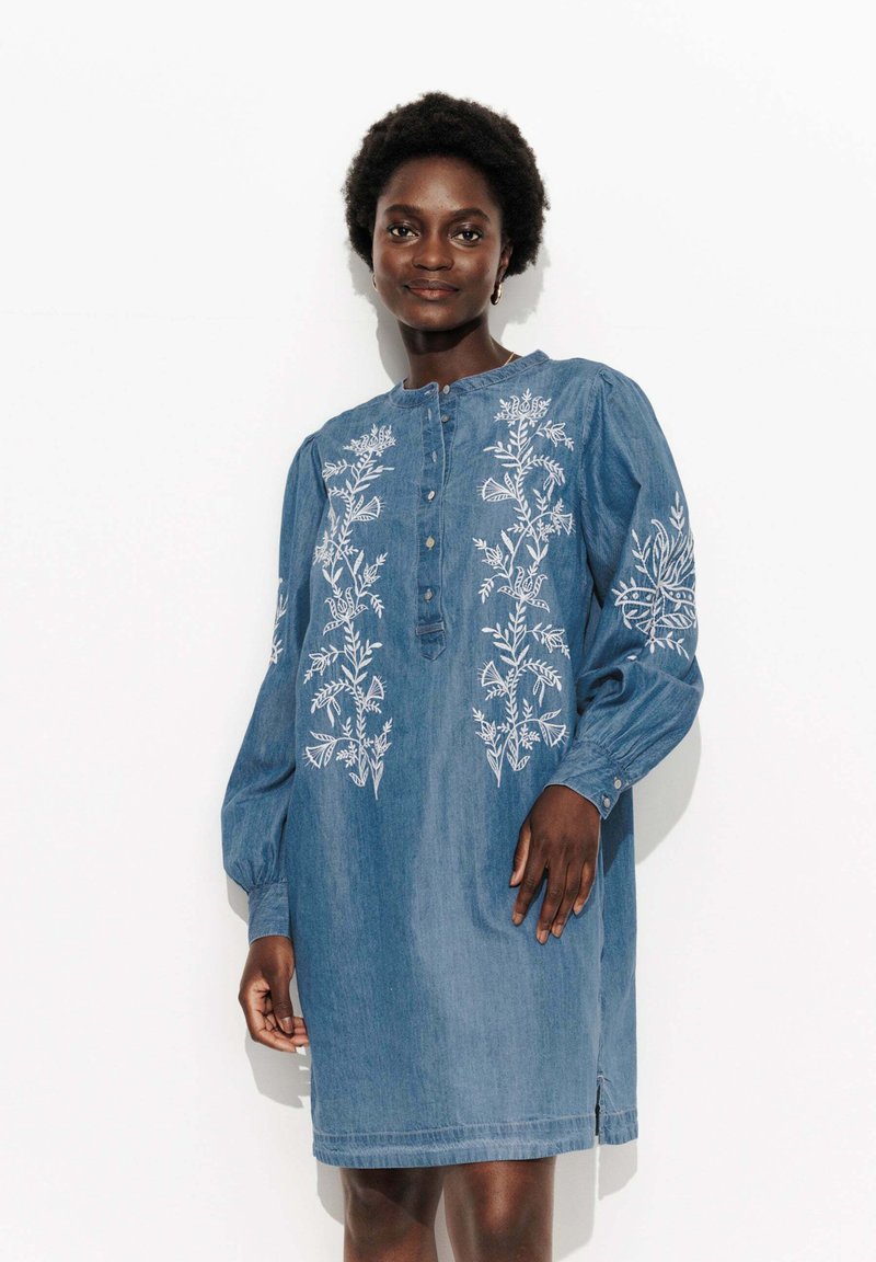 Robe en denim avec des manches longues, une patte de boutonnage et une broderie florale blanche complexe sur le devant et les manches.