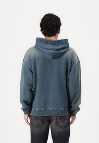 Hombre vestido con una sudadera con capucha azul desgastada y jeans gris oscuro, de pie frente a un fondo blanco liso.