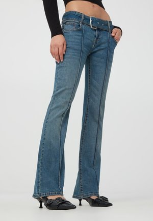 Jeans in denim blu a zampa d'elefante con cuciture a contrasto, cinque tasche e grande passante per cinta. Indossati con scarpe nere a punta.