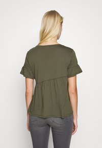 Olivově zelený top s krátkým rukávem a nařaseným pasem. Měkký jersey materiál, kulatý výstřih a volánkové lemování rukávů dodávají texturu a tvar.