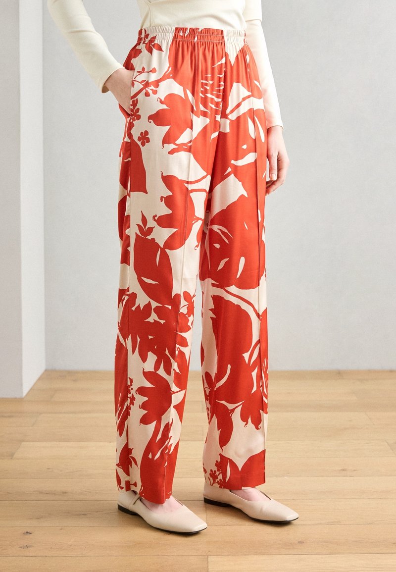 Femme portant un pantalon à jambes larges avec un grand imprimé floral rouge et crème, un haut blanc et des chaussures plates crème, debout sur un sol en bois.
