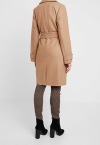 Long manteau couleur chameau en tissu doux, avec une taille ceinturée et des manches retroussées. Associé à un pantalon marron texturé et des bottines noires.