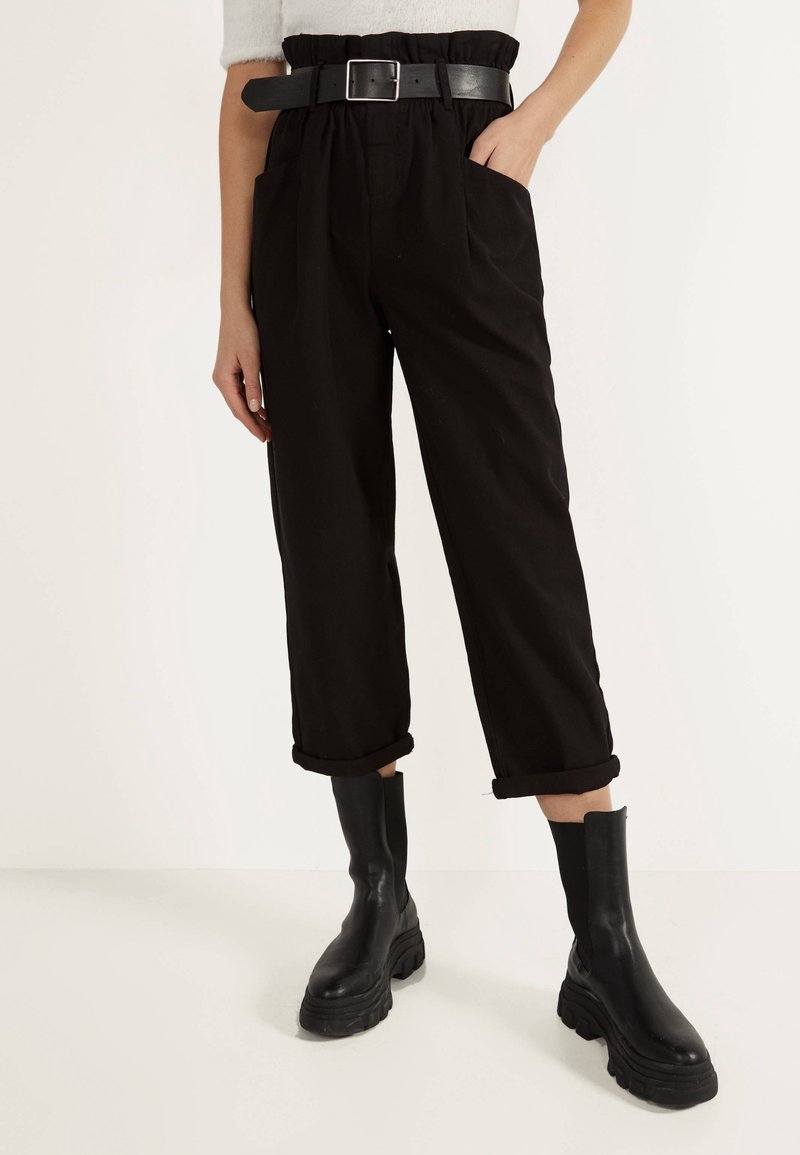 Pantalons noirs à ceinture, avec une coupe décontractée, présentant des ourlets retroussés et des poches latérales, associés à des bottes noires chunky.