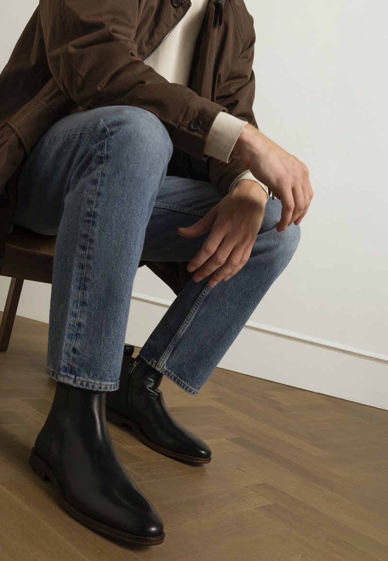 Schwarze Leder-Stiefeletten mit spitz zulaufender Zehe und minimaler Naht, kombiniert mit hellblauen Jeans und einem braunen Mantel, sitzend auf einem Holzstuhl.