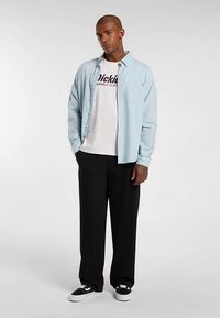 Vaaleansininen napitettava paita valkoisen t-paidan päällä, jossa on tummansiniset tekstit, mustat housut ja mustat kengät. Paidalla on rento istuvuus ja pitkähihaiset hihat.