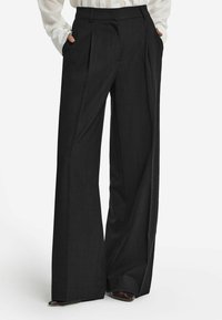 Svarta vida byxor i texturerat tyg. Har framfickor och veck, samt en skräddarsydd passform vid midjan.