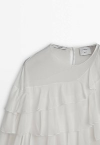 Blouse blanche avec des volants superposés sur le devant et les manches, encolure ronde et fermeture par bouton avec trou à l’arrière.