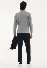 Jersey gris de punto, corte holgado, cuello redondo. Combinado con pantalones oscuros y zapatillas blancas. Sosteniendo una chaqueta oscura con la mano izquierda.