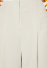 Pantaloni color crema realizzati in tessuto testurizzato, con pince frontali evidenti e un design su misura, caratterizzati da una finitura liscia.