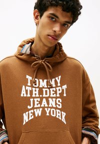 Hoodie marron avec le texte blanc "TOMMY ATH.DEP. JEANS NEW YORK". Doté d'une capuche avec cordon de serrage et d'un col à carreaux superposé. Matière douce et texturée.